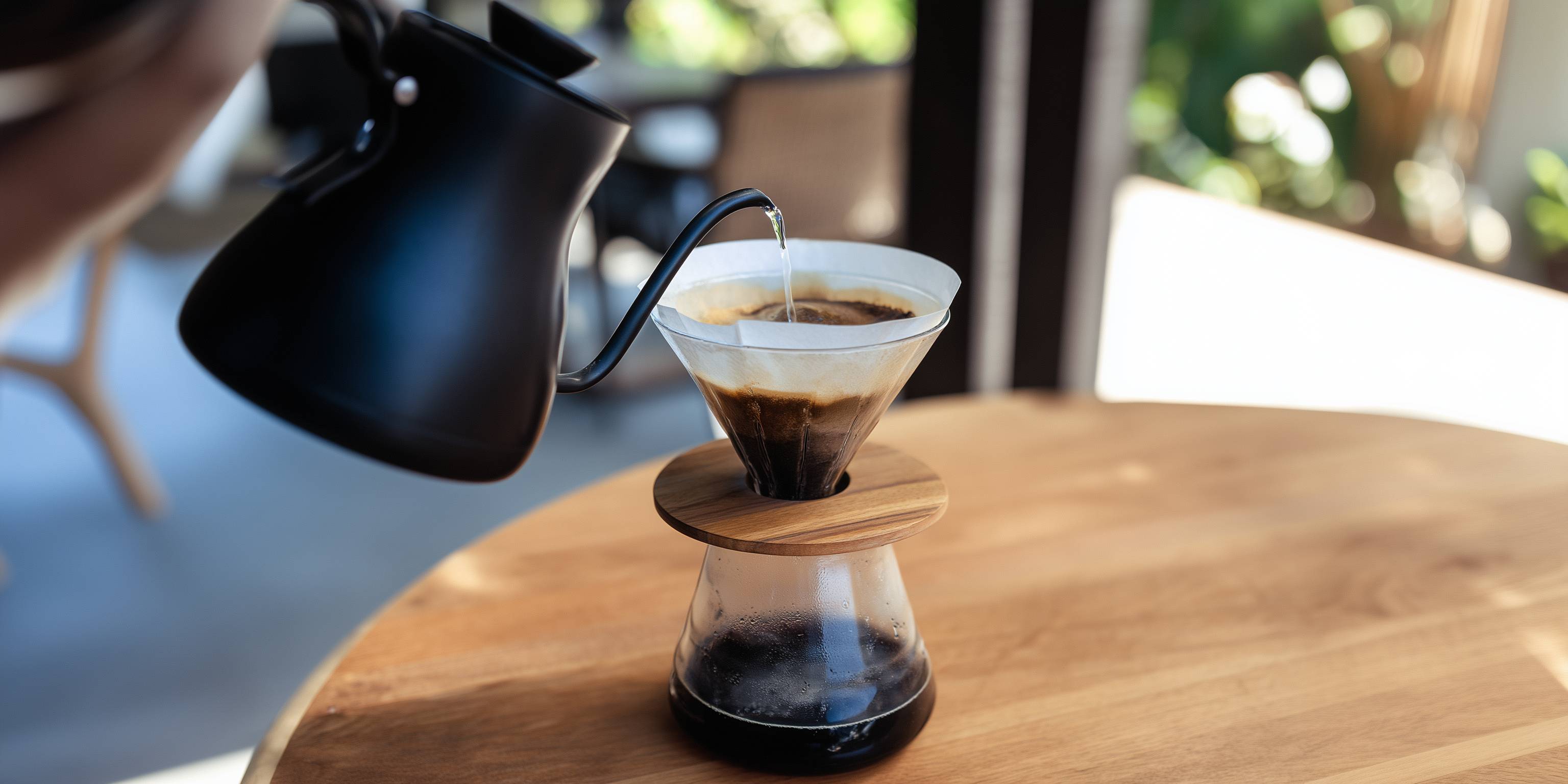 Handfilter-Kaffeezubereitung mit Kanne und Glasgefäß auf Holztisch.