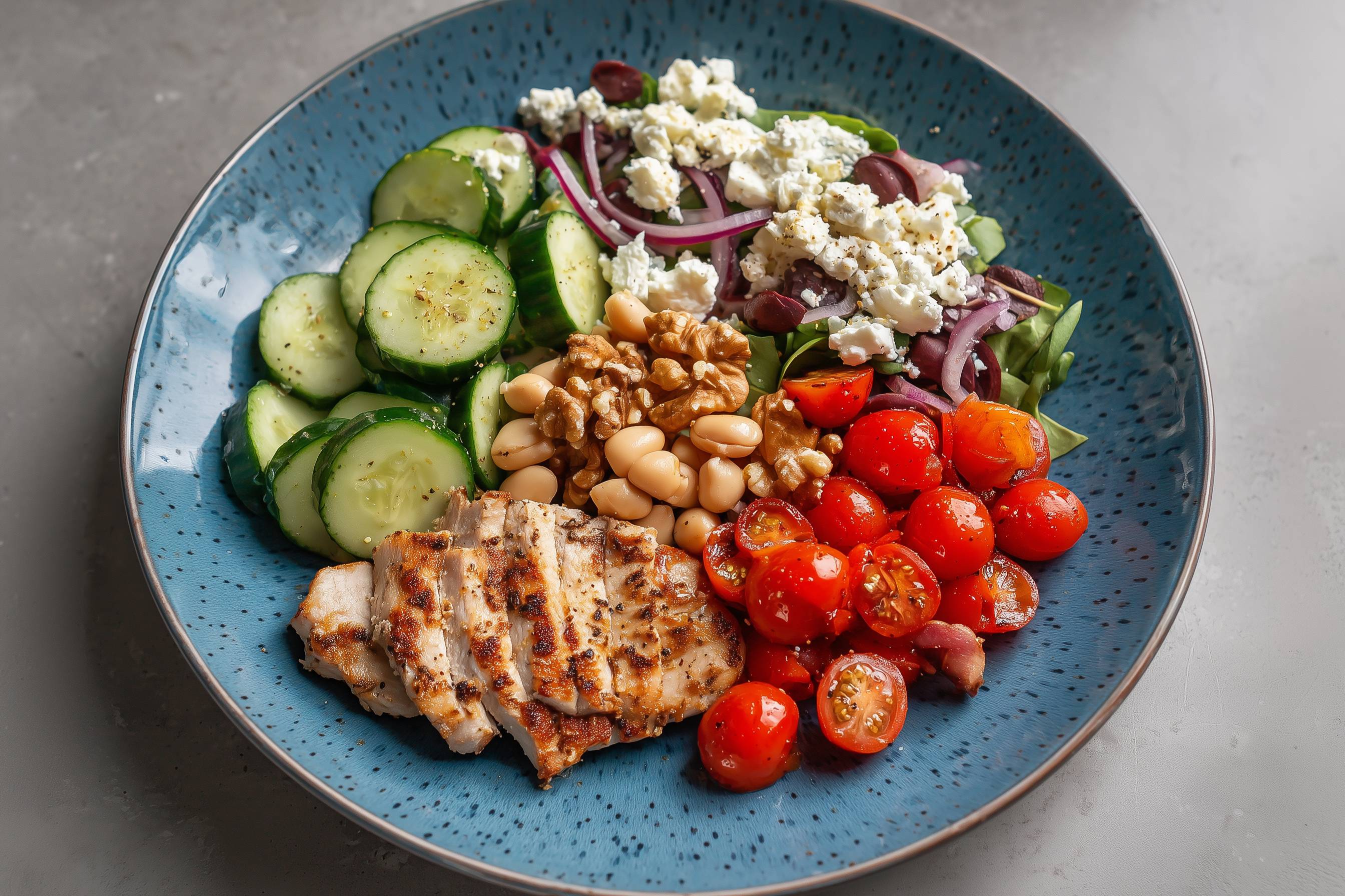 Ein Teller mit gegrilltem Hähnchen, Gurkenscheiben, Kirschtomaten, Bohnen, Walnüssen, Zwiebeln, Feta und Oliven.