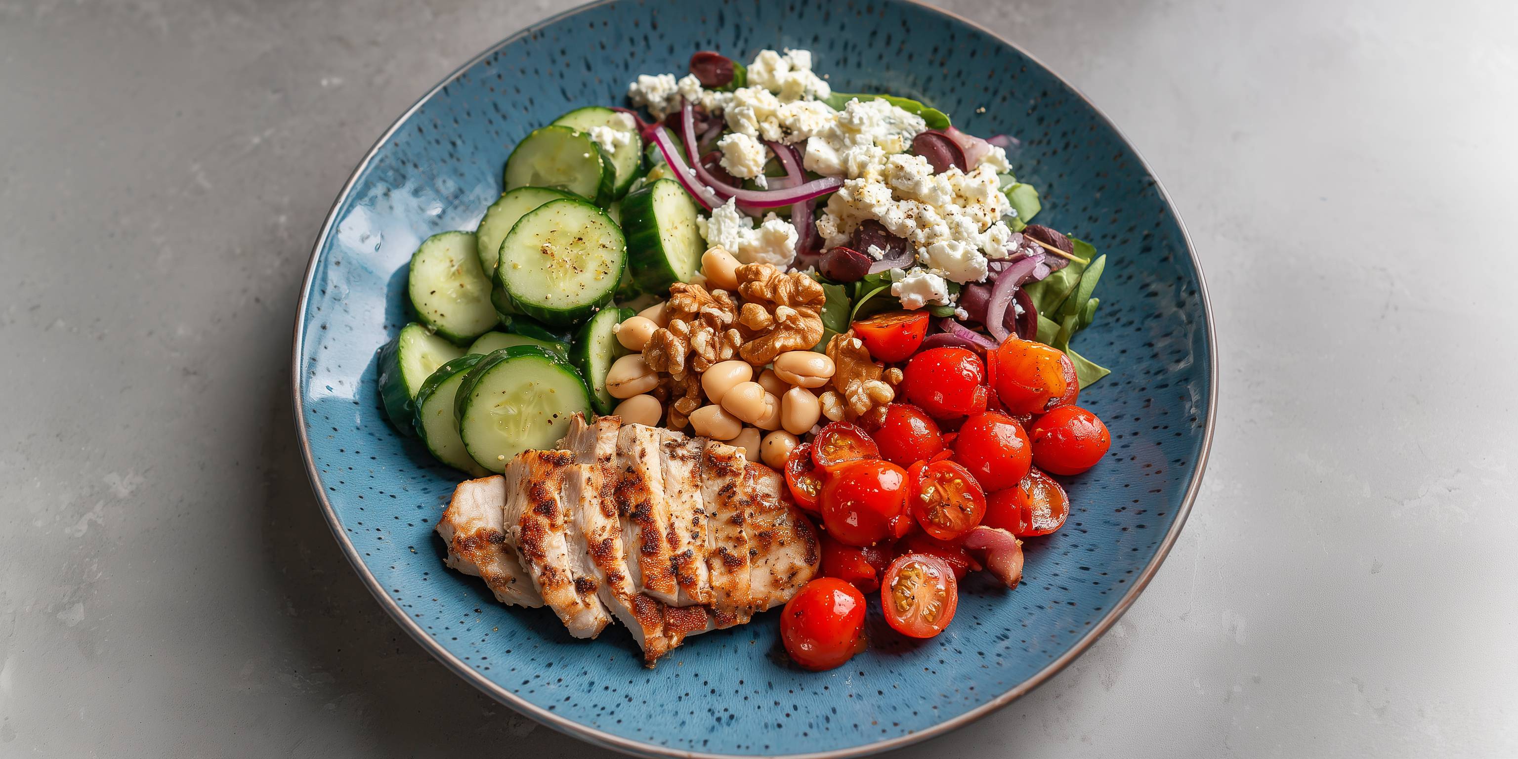 Ein Teller mit gegrilltem Hähnchen, Gurkenscheiben, Kirschtomaten, Bohnen, Walnüssen, Zwiebeln, Feta und Oliven.